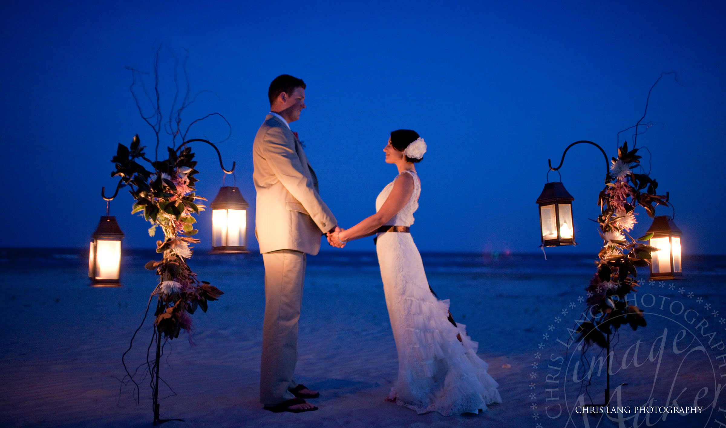 Wedding picture of a wedding couple holding hands at twilight in fron of some lanterns while on the beach after the beach wedding.  Destination wedding photographers