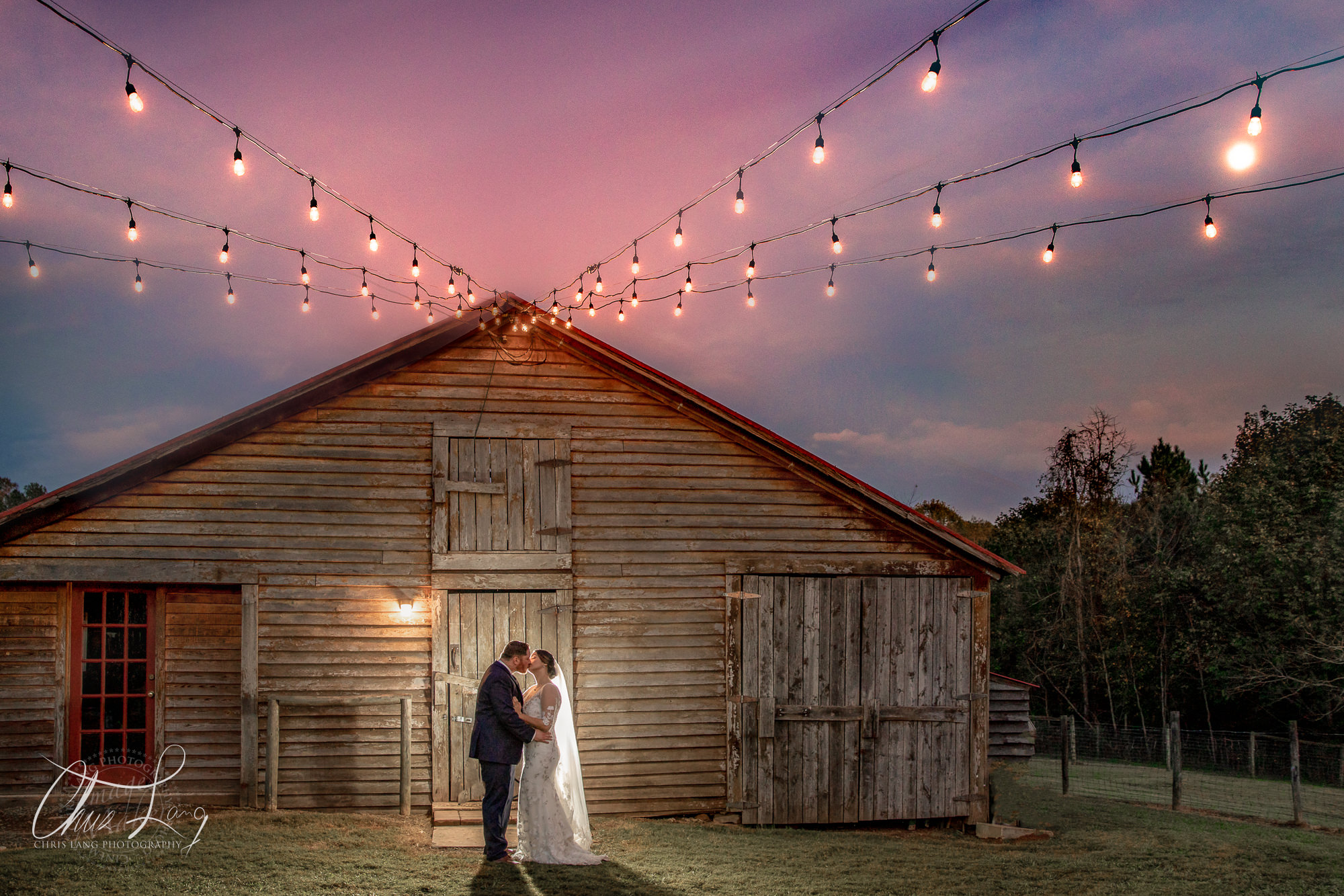wilmingotn nc wedding photographer - wedding photo  - creative wedding photography - photo of bride & groom  in fron a barn under edison lighting - twilight wedding photo